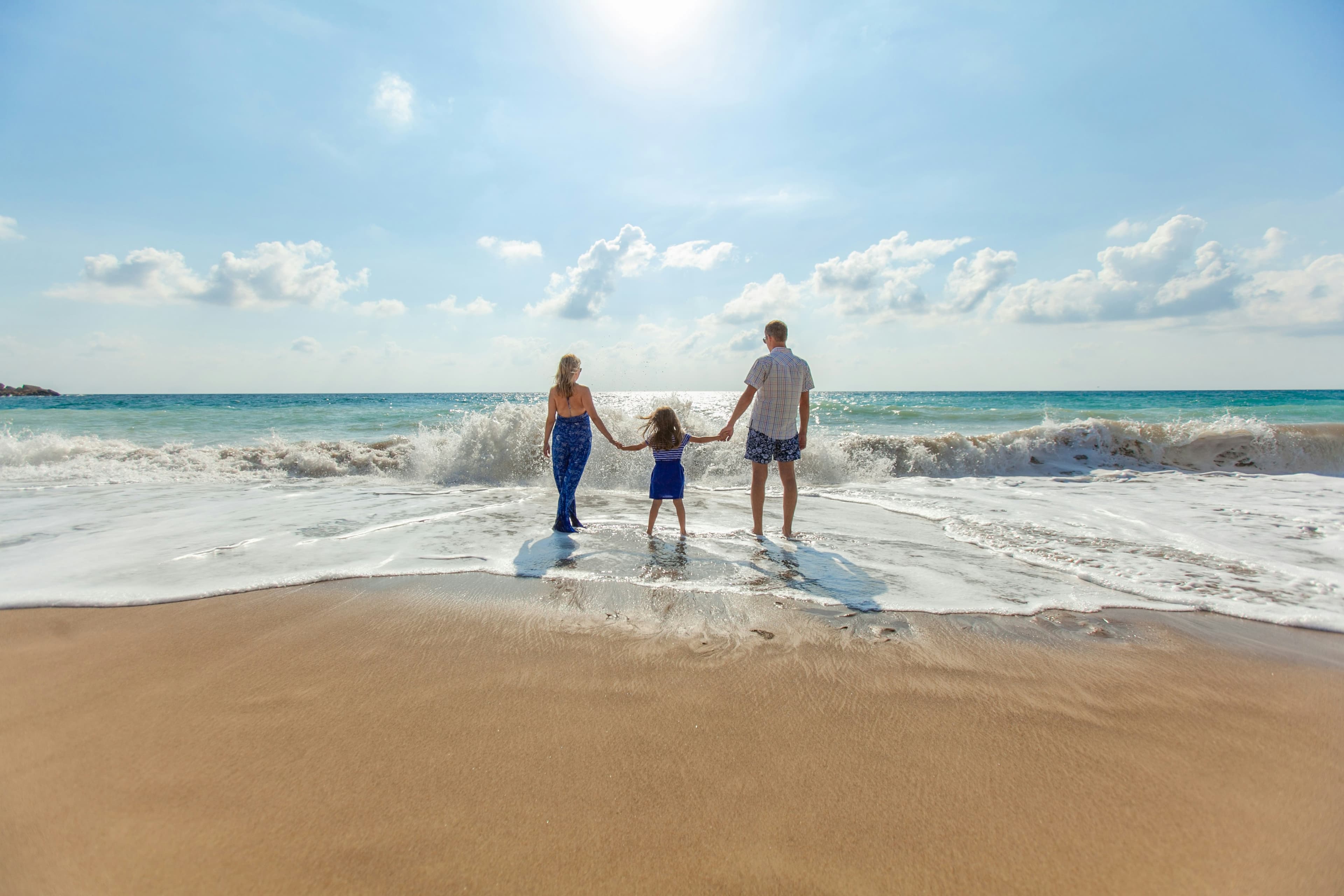 Parents and a child holding hands at the edge of the surf at the beach.
